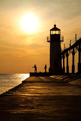 Silhouette of two men fishing off a lighthouse pier during sunset.
