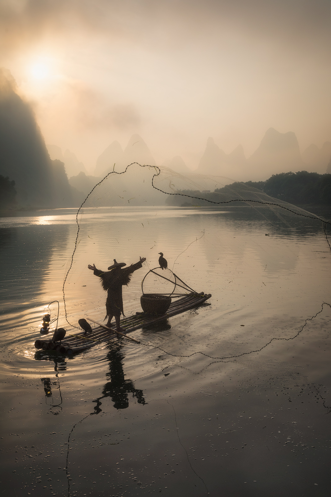 Chinese fisherman throwing his net just as the sun peaks around a karst mountain, creating a warm morning glow.