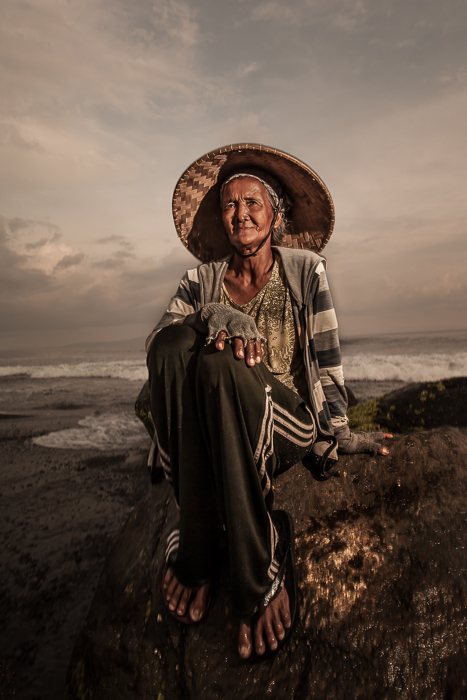 An elderly Indonesian woman sitting on a rock on the beach at sunset.