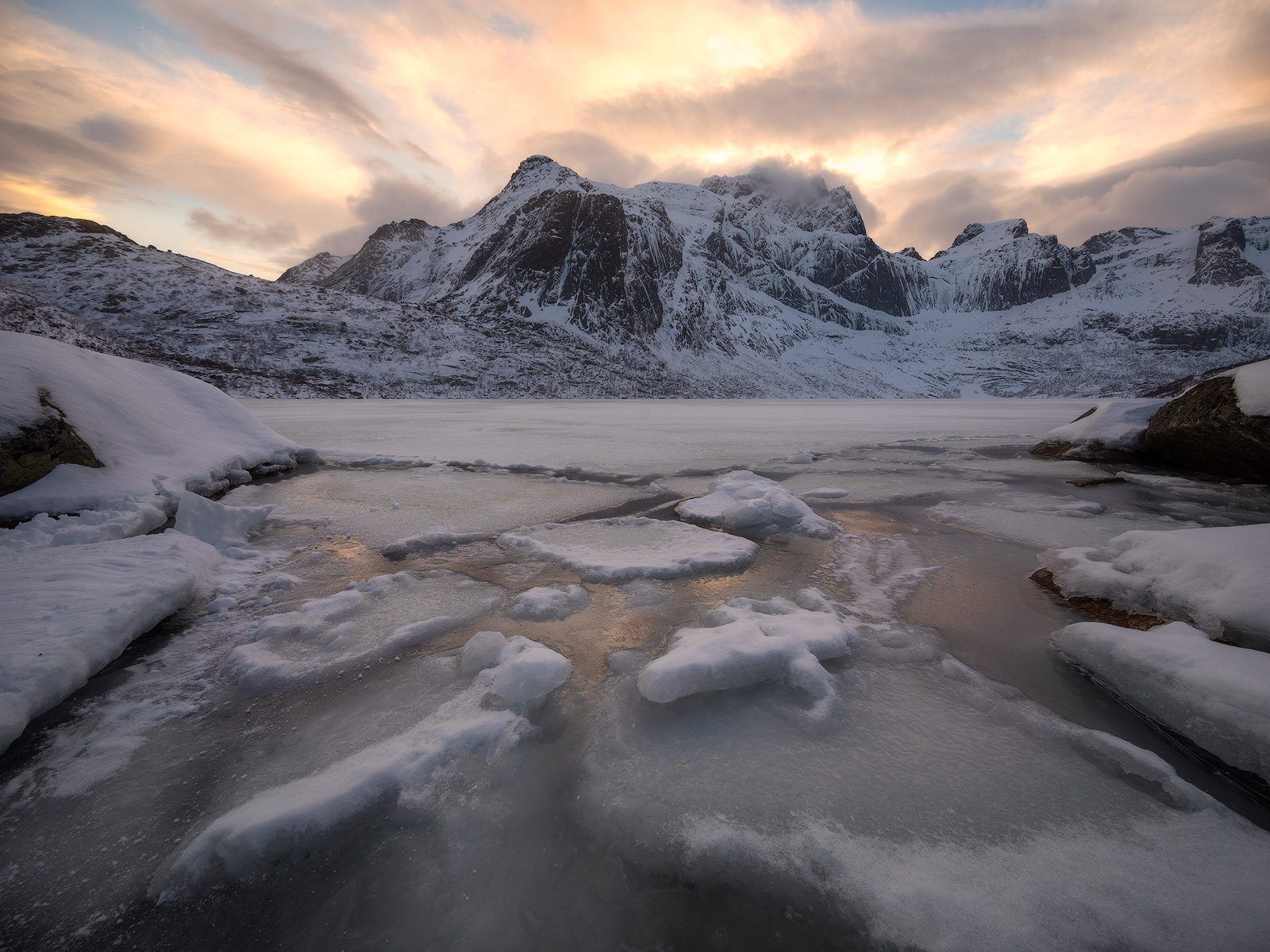 An icy lake forms leading lines to a snow covered mountain range.