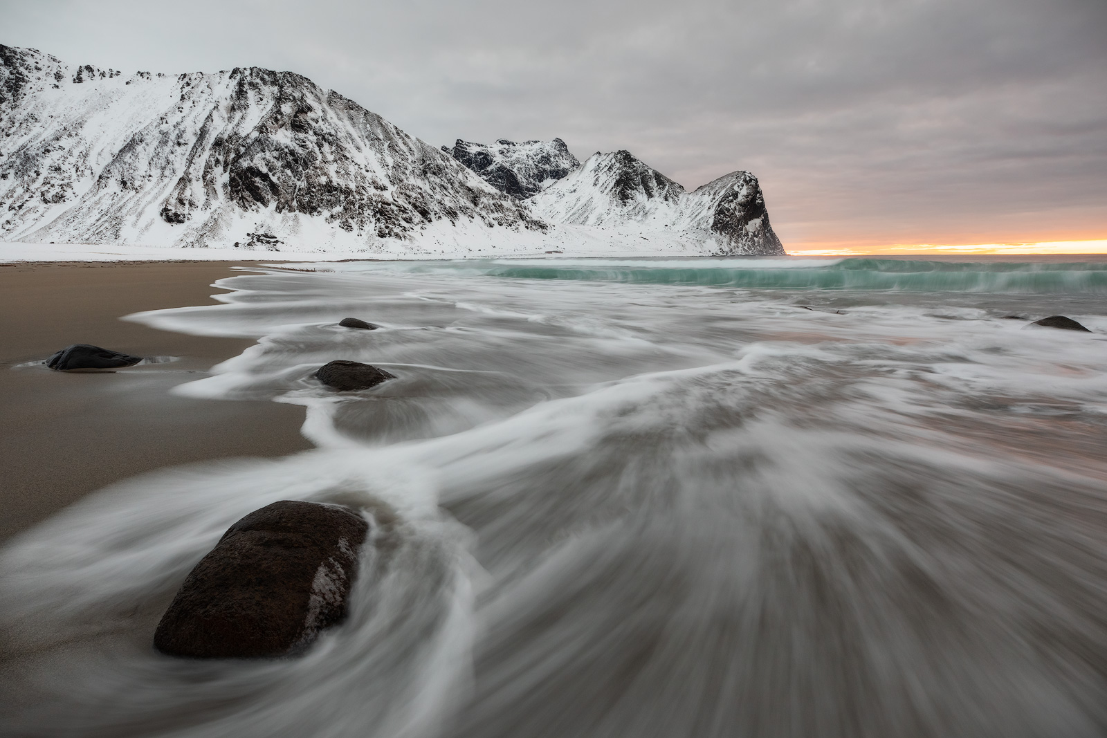 Waves hitting Unstad Beach as sunset peaks through a break in the clouds behind snow-capped mountains.