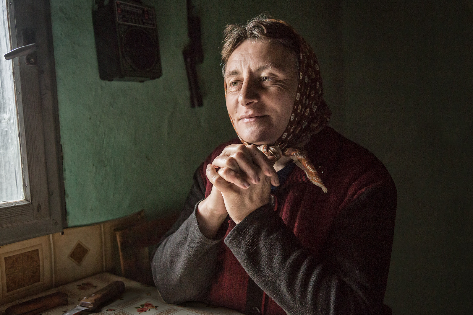 A rural Romanian woman sitting at her table is side-lit by window lighting.