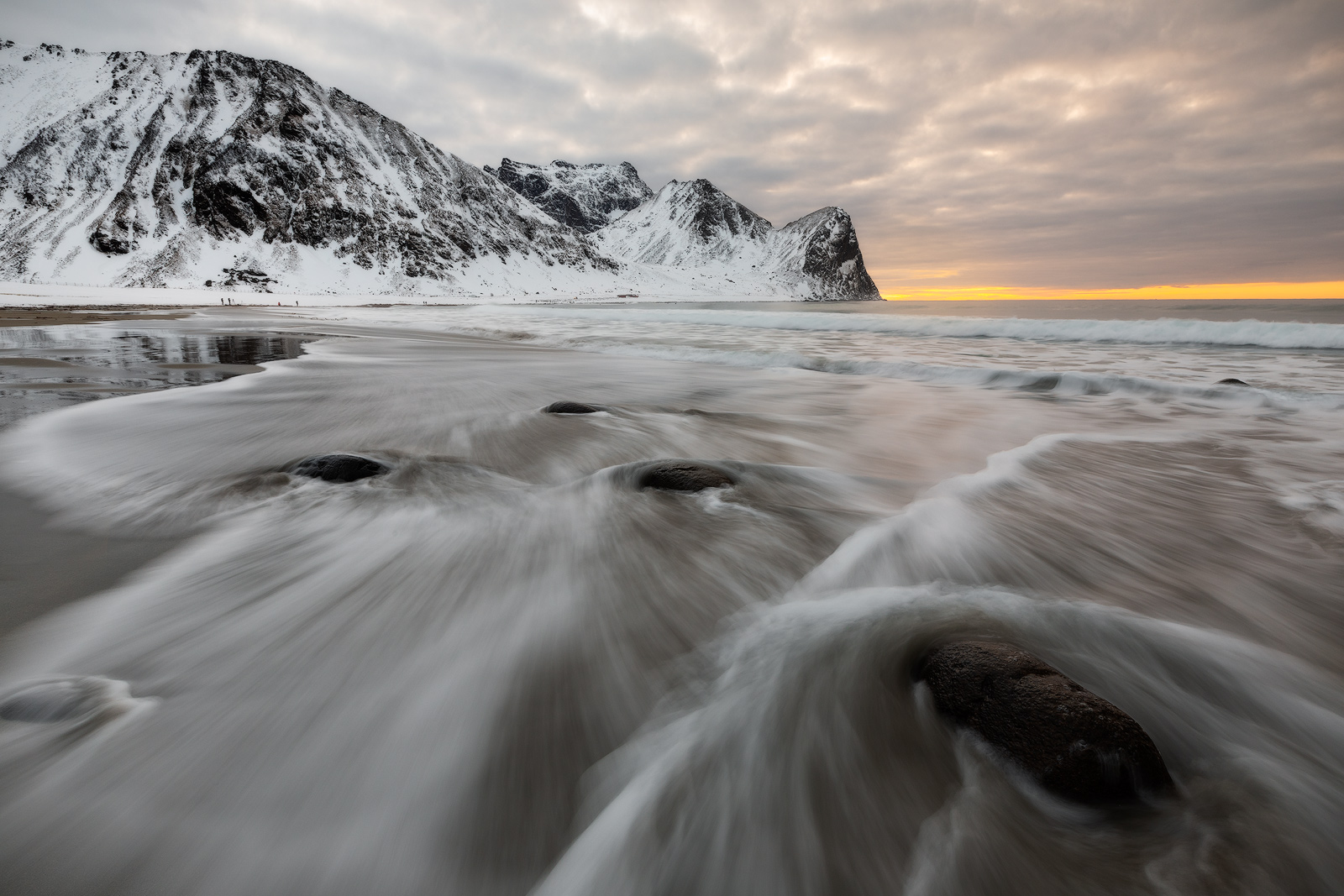 Snow-capped mountains line the beach while changing tide conditions create foreground leading lines.