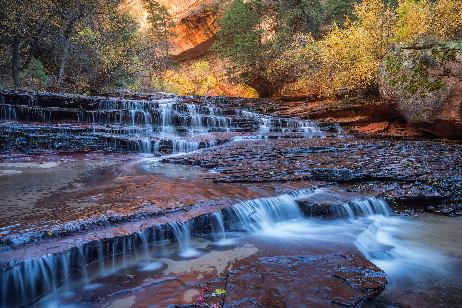 A trail interrupted by a beautiful waterfall surrounded by autumn leaves.